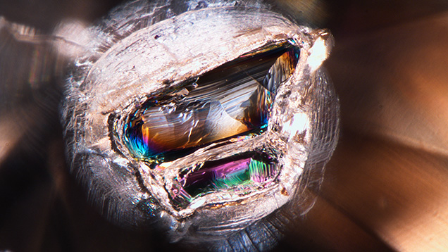 This iridescent ferropericlase crystal with a stress halo measuring approximately 0.7 mm was trapped inside a light brown diamond. Photomicrograph by Kyaw Soe Moe; field of view 1.58 mm.