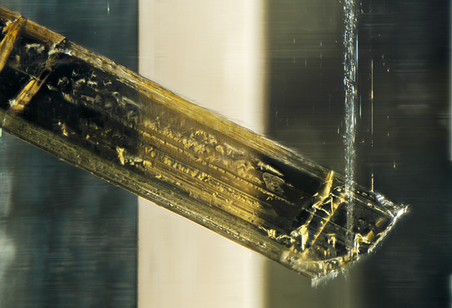 Morphological features of the tourmaline crystal included in the heliodor are visible; striations and triangular growth marks are present. A band of fluid inclusions is visible near the tip of the schorl tourmaline. Photomicrograph by Rhiana Elizabeth Henry; field of view 2.90 mm. Gift of Mark Mauthner, GIA Museum no. 37772.