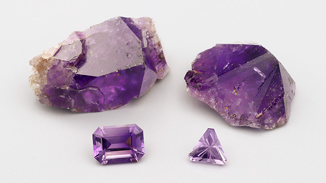 Figure 7. La Manche amethyst rough and faceted stones. The cut stone on the left (5.94 ct) shows diffuse color zoning. The one on the right (2.57 ct) is an example of light purple amethyst. Photo by Philippe Belley.