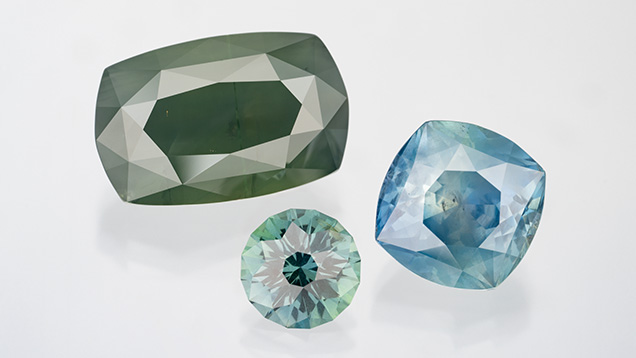 Figure 2. Three Montana sapphires showing a range of silkiness, from virtually none (2.80 ct green round cut by Craig Oliveira) to moderate (10.33 ct blue cushion cut by Victor Tuzlukov) to strong (21.20 ct hazy green “silk bomb” cut by Tuzlukov). Photo by Robert Weldon; courtesy of Obsessed Over Gems.