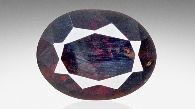 Figure 1. A 0.85 ct oval modified brilliant titanoholtite. Although its bodycolor was dark orangy brown, a bluish haze was visible from certain angles due to light scattering off polish lines as well as internal colorless graining. Photo by Adriana Robinson.